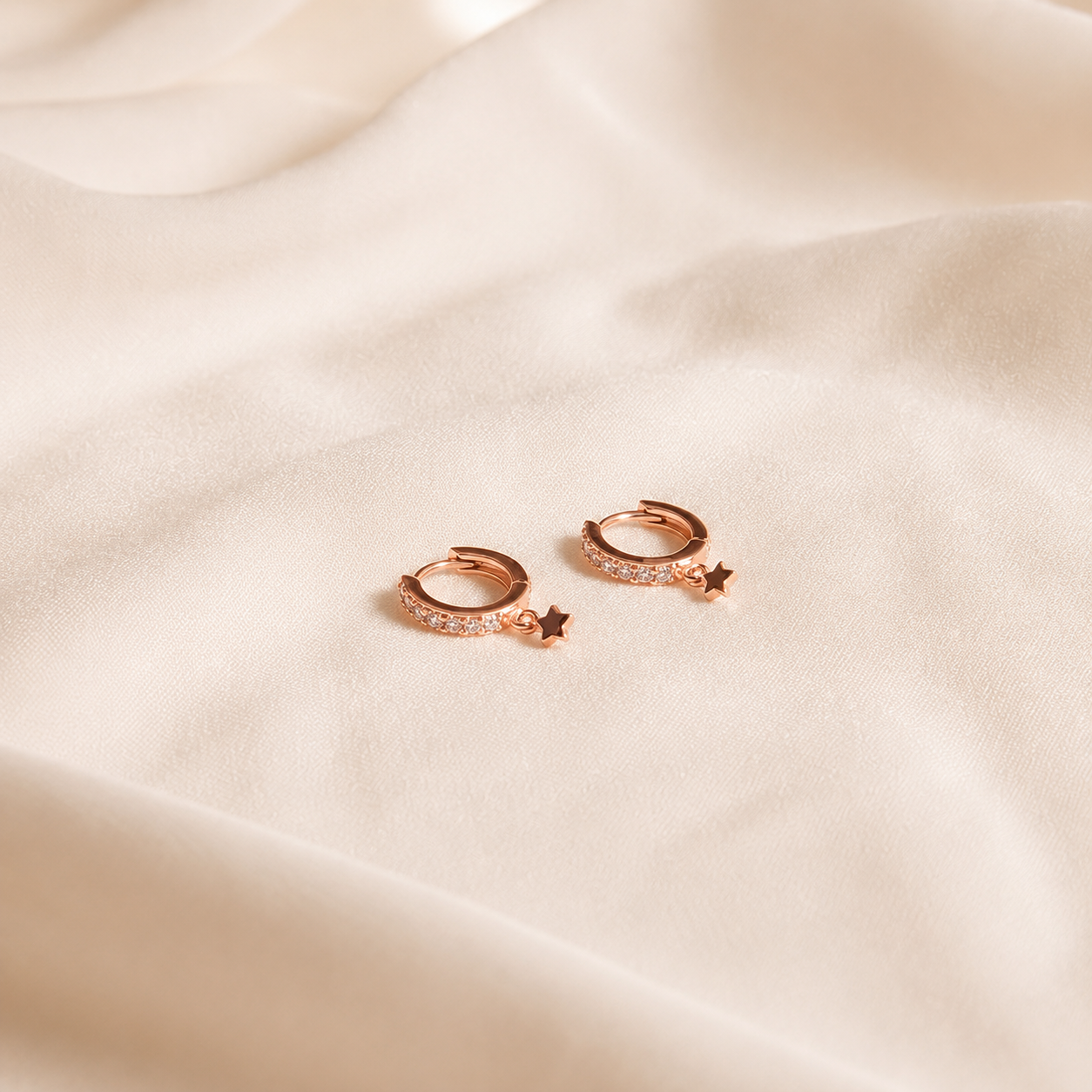 A pair of small rose gold huggie hoop earrings with tiny pavé-set stones and dangling star charms, resting on draped cream-colored fabric.